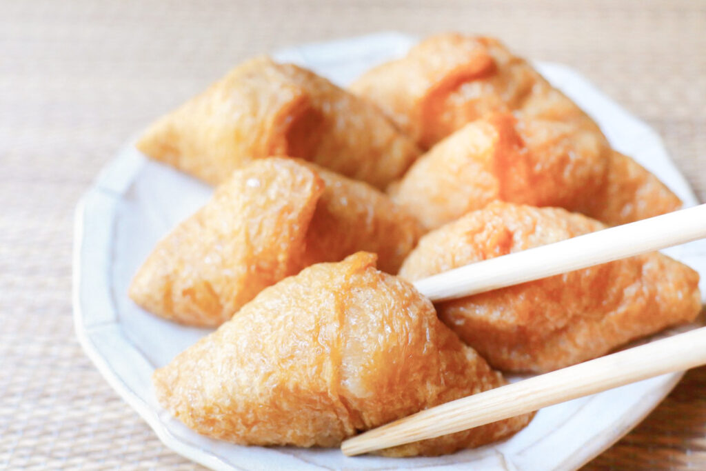 Six triangle-shaped inari sushi on a white plate sitting on a textured beige placemat. The piece closest to camera is about to be picked up by a pair of light-colored wooden chopsticks from the right of screen.
