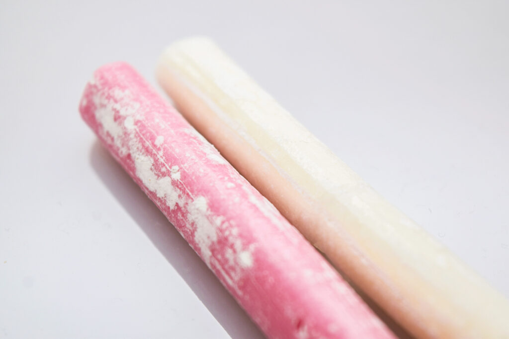Two long cylindrical sticks of chitose ame candy - one pink (red) and one white - lay on a white surface. It is a close-up shot with only part of the chitose ame in view.