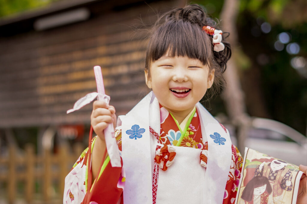 A three-year-old girl wears a red kimono with a white 'hifu' vest at a shrine in celebration of Shichi go san. She faces the camera with such a huge grin that her eyes appear closed. She holds up a stick of red (pink) chitose ame in her right hand. The chitose ame bag can be seen in her left hand.