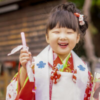 A three-year-old girl wears a red kimono with a white 'hifu' vest at a shrine in celebration of Shichi go san. She faces the camera with such a huge grin that her eyes appear closed. She holds up a stick of red (pink) chitose ame in her right hand. The chitose ame bag can be seen in her left hand.