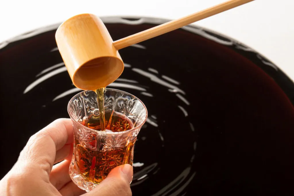 A large open barrel of black Japanese vinegar. The shot has been taken from the point of view of someone pouring some into a small glass cup with a bamboo ladle.