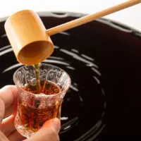 A large open barrel of black Japanese vinegar. The shot has been taken from the point of view of someone pouring some into a small glass cup with a bamboo ladle.