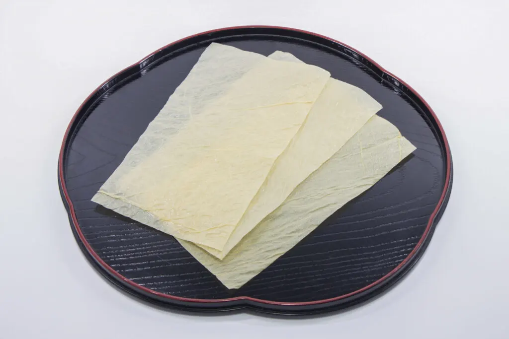 An example of yuba (tofu skin) from Kyoto. There are three very thin rectangular pieces in dried form fanned out on a black lacquered wooden serving board with rounded edges and a red trim. The pieces of tofu skin are so thin that you are able to see the other pieces and board through them. 