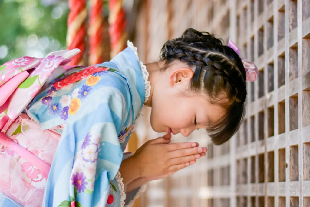 A seven-year-old girl in a light blue kimono with a floral pattern and a light pink obi sash bows in prayer at a shrine. Her hair is braided in an intricate up do with a light pink hair accessory.