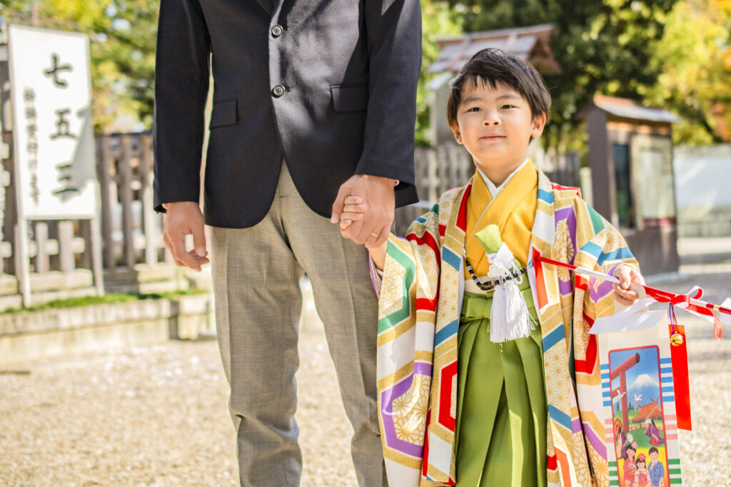 A five-year-old boy (right) in traditional hakama pants and jacket holds hands with his father (left) wearing Western-style pants and a suit jacket (we can only see the father until chest height). In his free hand, the boy holds a bag of chitose ame candy and a 'hamaya' arrow lucky charm for dispelling demons and bad luck.