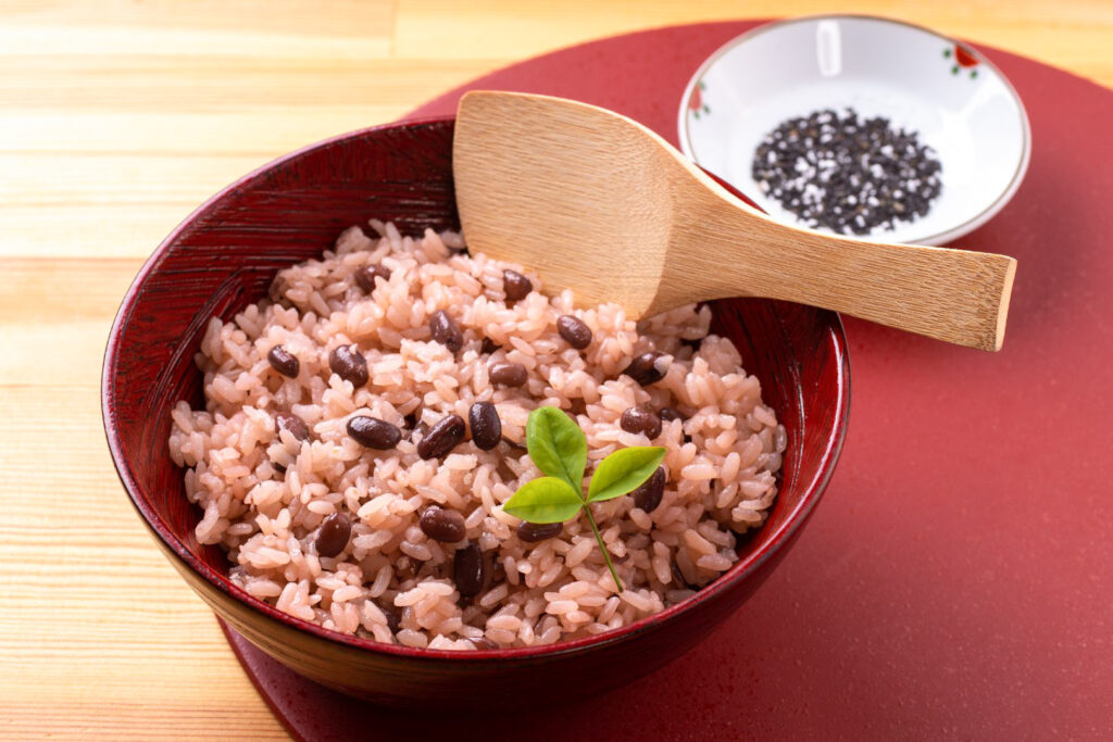 Shichi go san food: A red bowl filled with 'sekihan', glutinous rice with red azuki beans that turn the rice a reddish color and is eaten on celebratory occasions in Japan. The bowl has a leaf garnish and a wooden paddle for serving. Behind it is a small white bowl of black and white 'goma' (sesame seeds) commonly sprinkled on the dish.