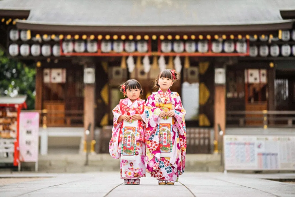 Two girls, one three and the other seven, celebrate Shichi go san in floral patterned white, pink and red kimono in front of a shrine. They are both standing side by side facing the camera, have red flowers in their pinned up hair, and are holding chitose ame candy bags with both hands in front.