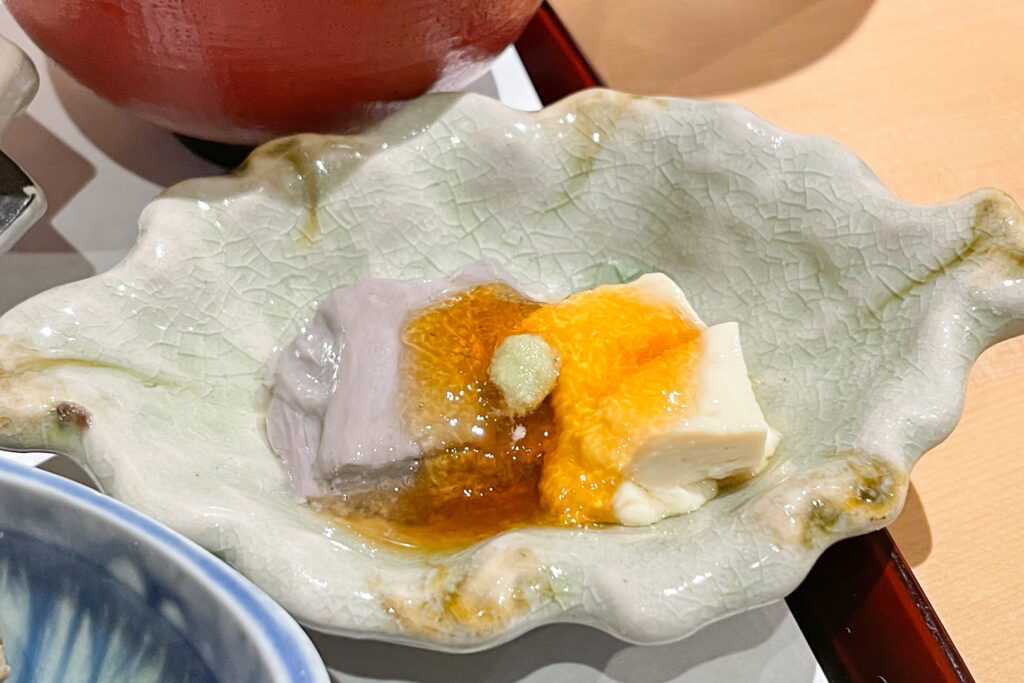 A close-up of one of the dishes in the aforementioned yuba lunch set. It has regular tofu skin as well as yuba made from black soybean, giving it a light purple-grey color. It is covered in an almost jello-like yellow sauce with a small dollop of wasabi in the center.