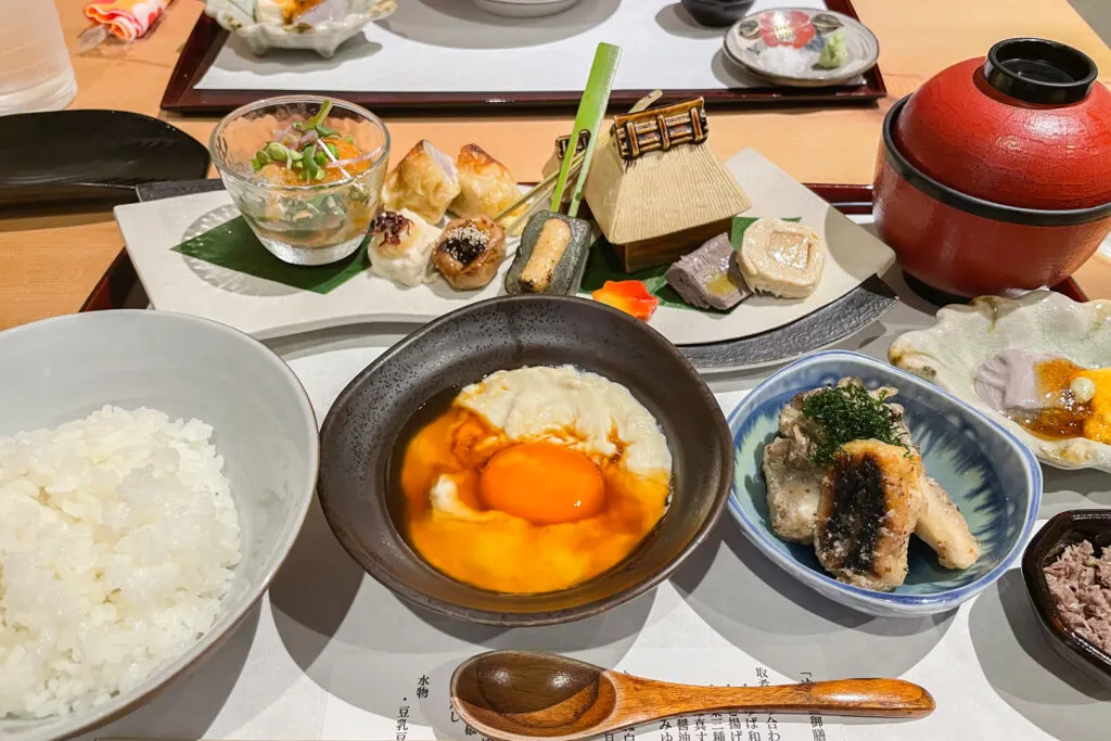 A lunch set at a restaurant in Kyoto with a variety of yuba dishes, including one with raw egg and soy sauce on top. There is also a bowl of white rice and a bowl of what is presumable miso soup with the lid still on.