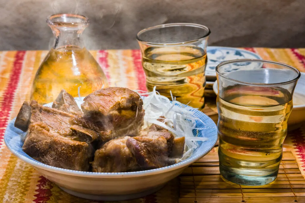 A clear glass vessel filled with awamori with a golden hue sits on a red, yellow and mustard striped tablecloth along with two full glasses of awamori and a plate of stewed 'sōki' Okinawan spare ribs.