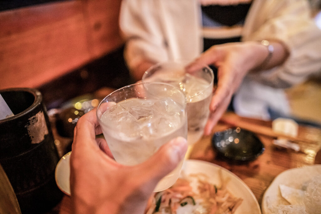 Two hands are bringing their glasses of iced and cut awamori together in a toast across a small restaurant table with some dishes on the table in Okinawa.