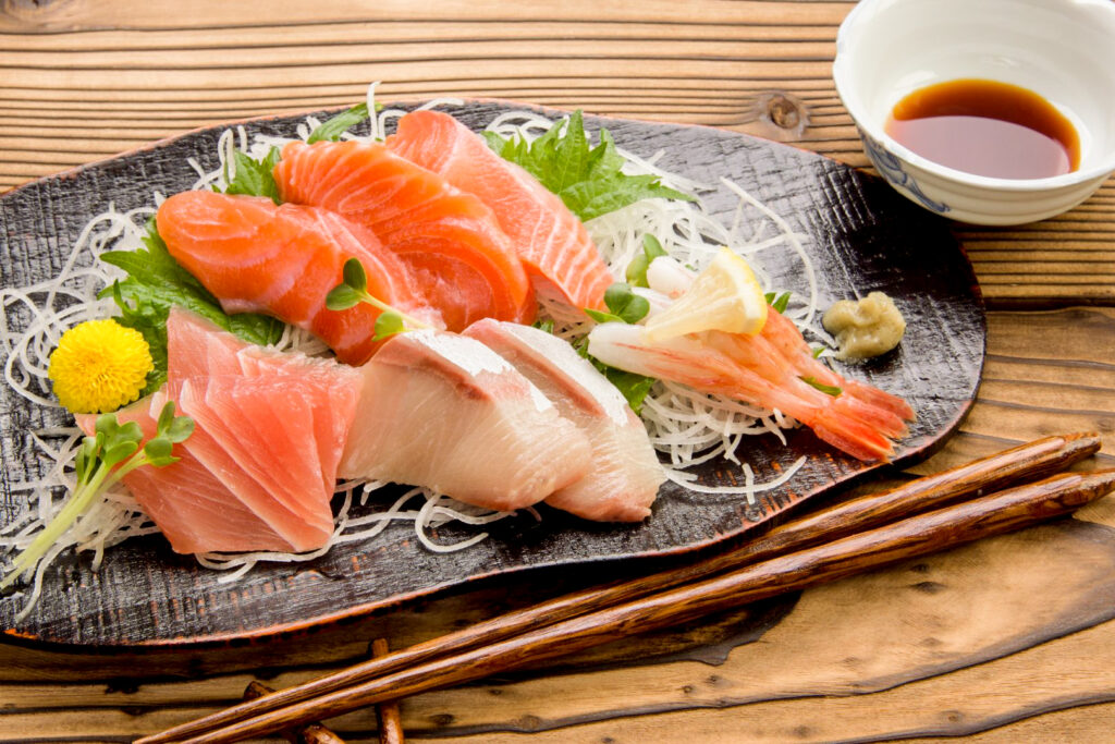 Various slices of raw fish on a flat dark ceramic plate, including salmon and prawns. They sit on some shiso leaves and a bed of daikon radish strings. Other sashimi garnishes neatly arranged around the fish include a chrysanthemum flower and some green shiso sprouts. To the side of the plate is a dollop of what appears to be a wasabi-type condiment and next to it a small white ceramic bowl with some soy sauce in it. A pair of wooden chopsticks has been placed in front of the plate.