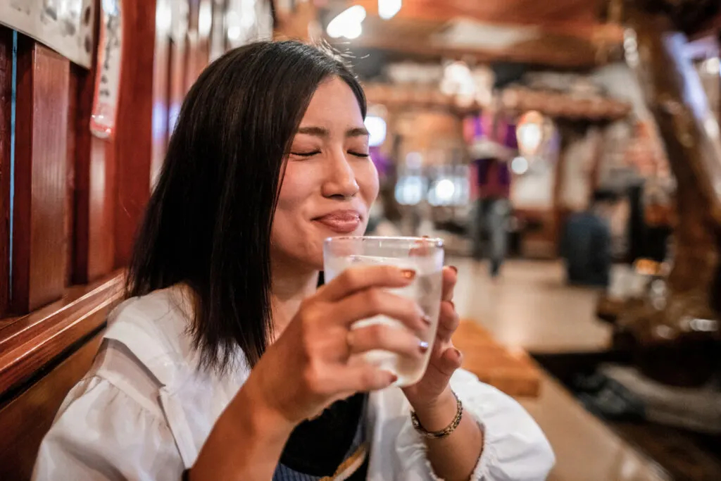 A Japanese woman with hair down to her collarbone and wearing a white jacket with a black top has her eyes closed and a satisfied smile on her face after taking a sip of an iced awamori cocktail. She is seated at a table by the wall with her back facing the wall. She appears to be in a drinking establishment.