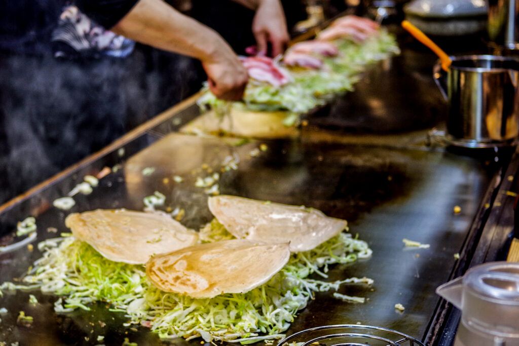 A large hot plate with three Hiroshima okonomiyaki in the foreground that have already been flipped. The cabbage and cooked batter disk on top can be seen. While in the background, a person is about to flip the first of five Hiroshima okonomiyaki lined up in a row.