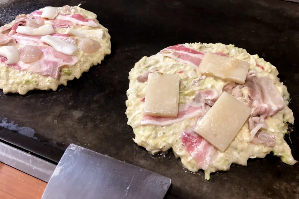 Two Kansai-style okonomiyaki being cooked on a hot plate. They have not been flipped yet so the batter is still wet on top. The one on the left is topped with sliced pork, scallops and squid, while the one on the right has sliced pork and mochi.