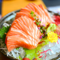 Perfectly arranged slices of raw salmon atop a bed of ice and daikon radish strings in a shallow dark ceramic bowl-plate. Other sashimi garnishes can also be seen such as shiso leaf, shiso flowers, a chrysanthemum flower, and red and green seaweed.