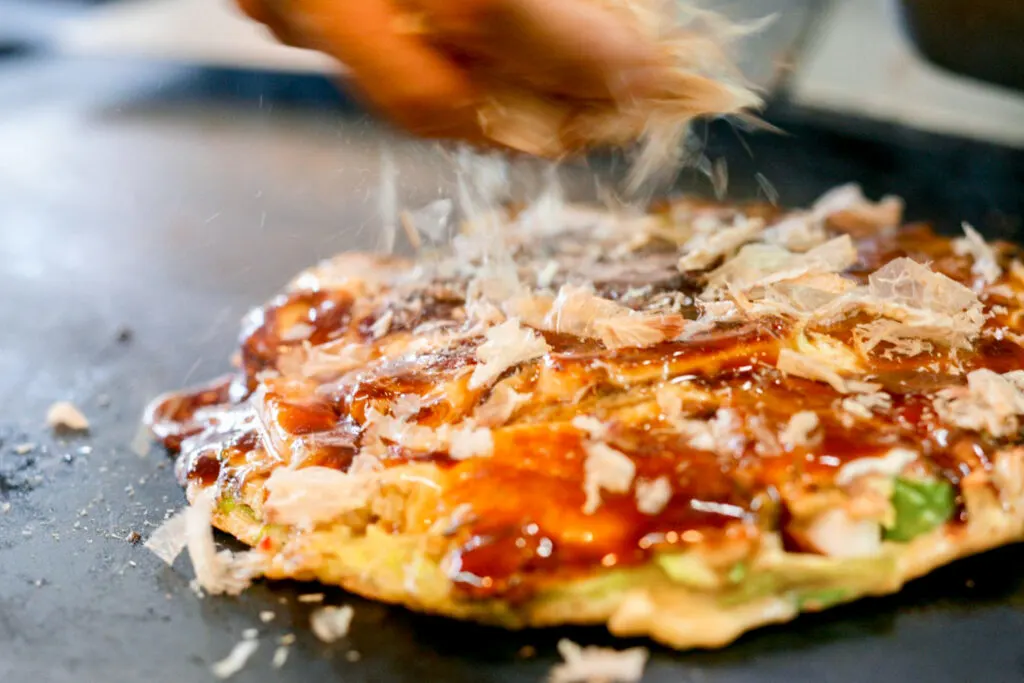 A Kansai-style okonomiyaki, also known as Osaka-style okonomiyaki, on a hot plate. It has been topped with brown okonomiyaki sauce and a hand can be seen sprinkling dried bonito flakes (katsuobushi) on top.
