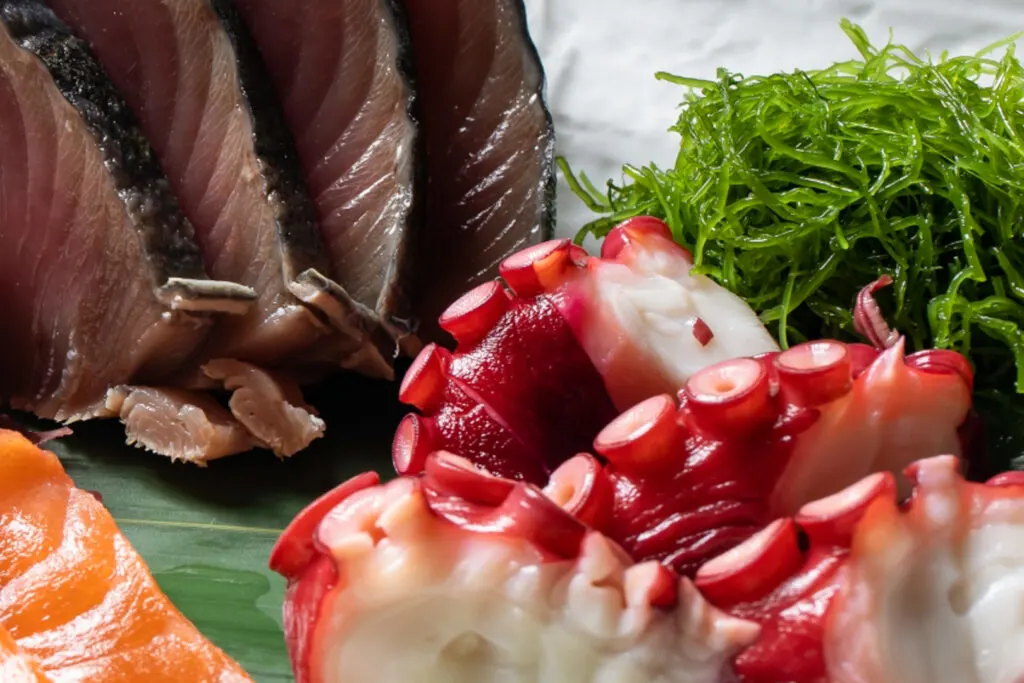 A close-up shot of a pile of stringy cooked green ogonori seaweed as part of a sashimi presentation. Octopus sits prominently in front of it.