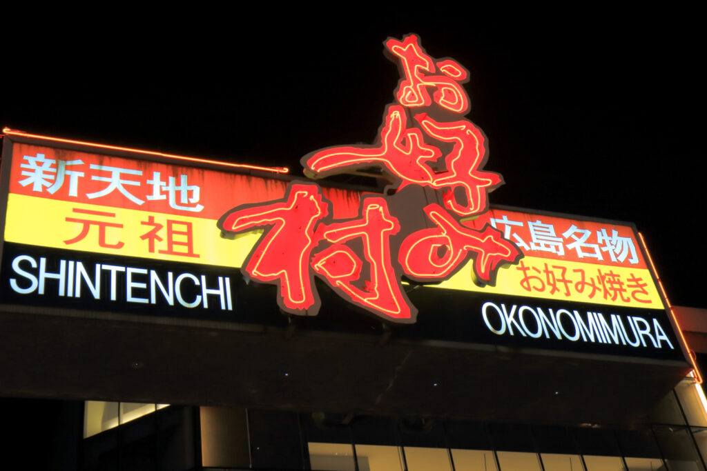 The lit-up red, yellow, black and white Okonomimura sign in Hiroshima at night. Down the center are large Japanese characters spelling Okonomimura.