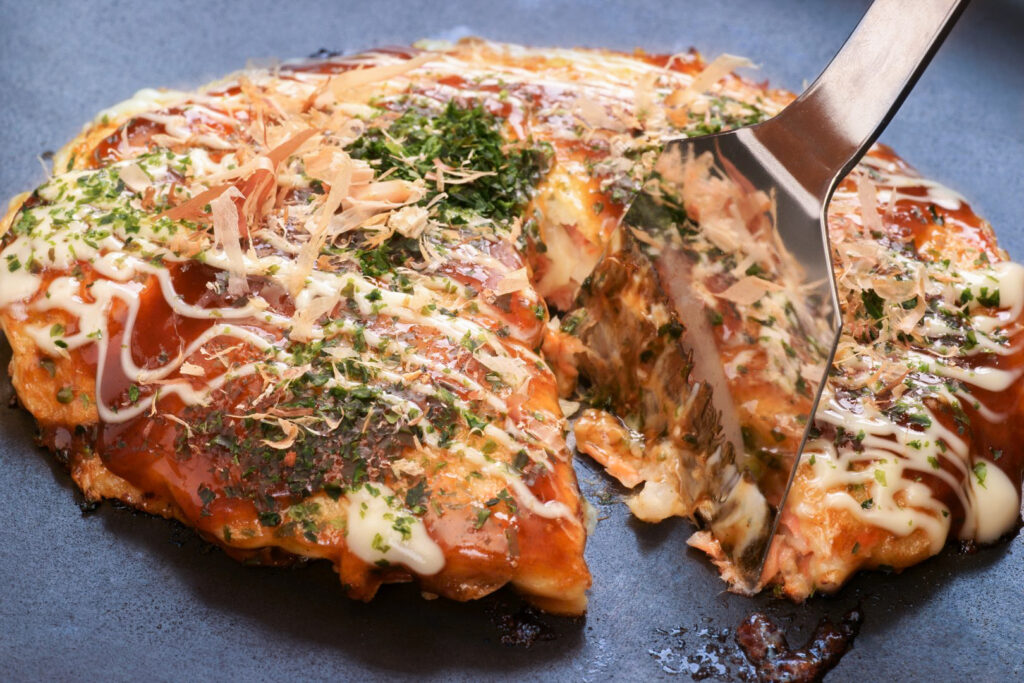 A wedge is being cut out of an Osaka-style okonomiyaki using a 'hera' spatula.