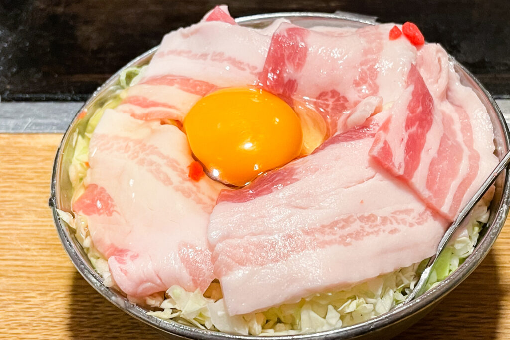 The raw ingredients for Osaka okonomiyaki in a silver metal mixing bowl. Shredded cabbage, slices of pork and a cracked raw egg can be seen on top.   