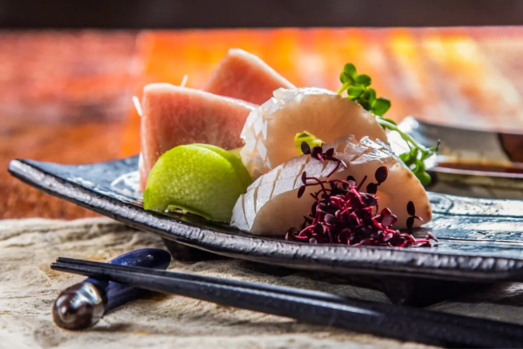 A side-on shot of an artfully presented plate of sashimi. The raw fish slices have been arranged standing on their sides, so the dish has a little height to it. Some red shiso sprouts can be seen at the front and some green daikon sprouts can be seen popplng up from the back. Although mostly obscured, some daikon radish strings can also be seen from the left and there is also what appears to be some lime at the front. Dark colored wooden chopsticks sit on a blue ceramic chopstick rest in front of the plate.