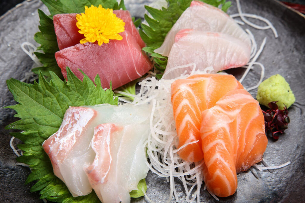 Various slices of sashimi atop daikon radish strings and numerous shiso leaves. A yellow chrysanthemum flower, red shiso sprouts and wasabi are also arranged on the plate.