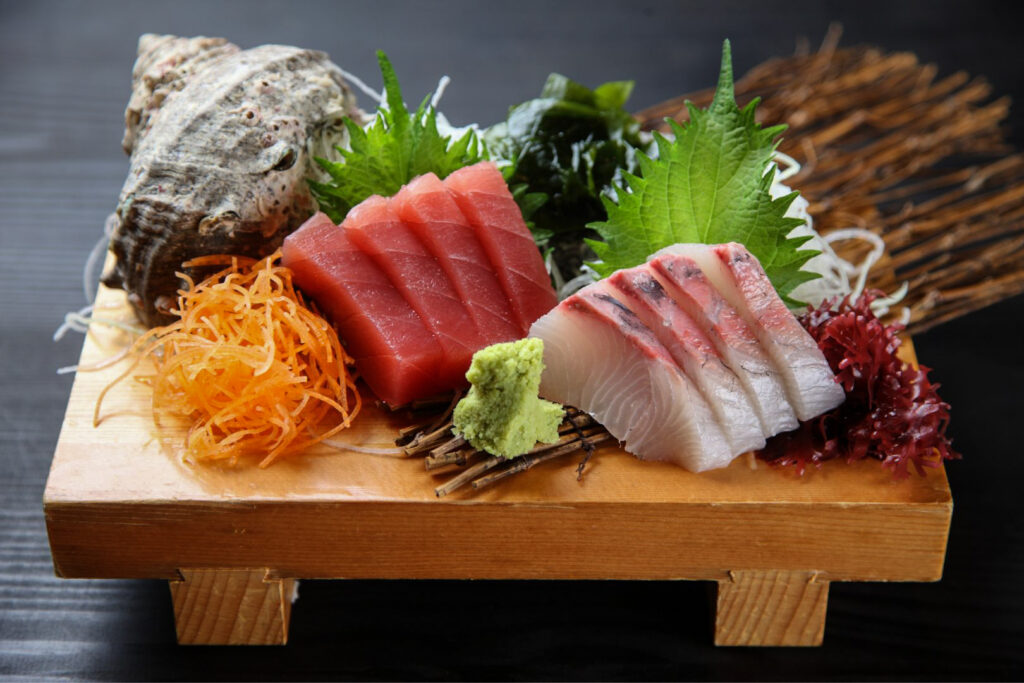 A sashimi presentation on a wooden board with legs that raise it a little off the tabletop. The slices of fish have been arranged on their sides ("standing") and around the fish are various sashimi garnishes including red tosakanori seaweed, shiso leaf, wakame seaweed, daikon radish strings, carrot strings and wasabi.