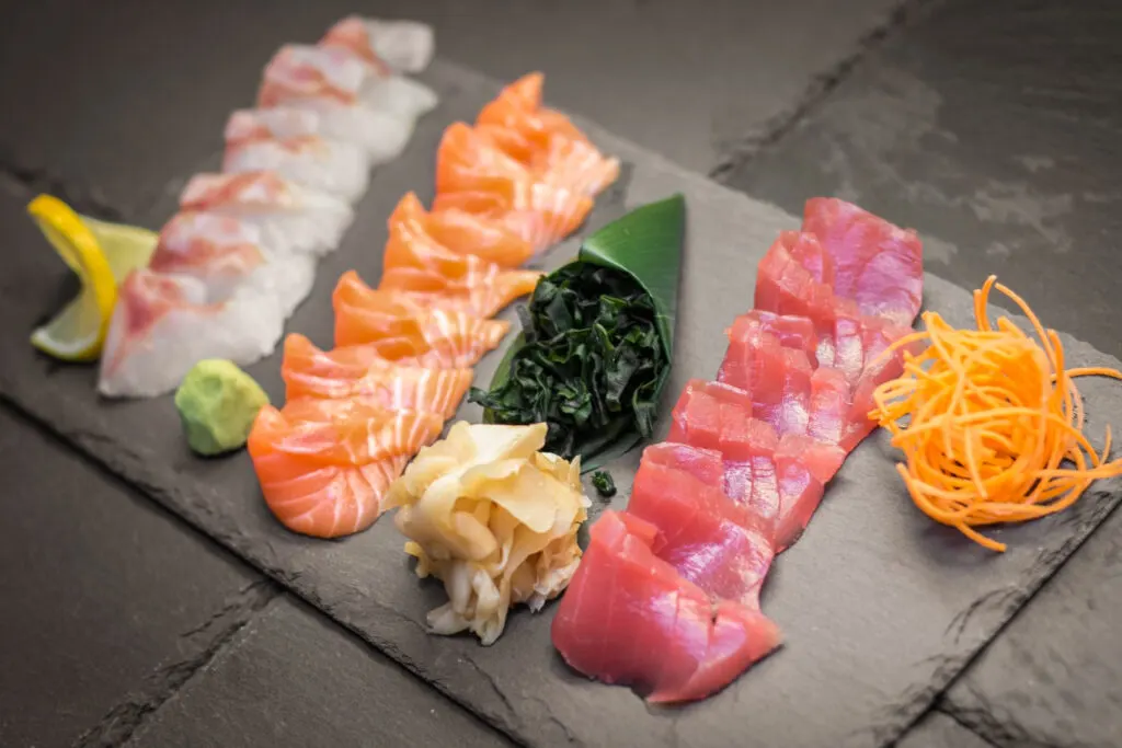 Various slices of raw fish lined up in rows on a dark slate board. The various sashimi garnishes arranged on the board include wakame seaweed, pickled ginger, carrot strings, wasabi and lemon.