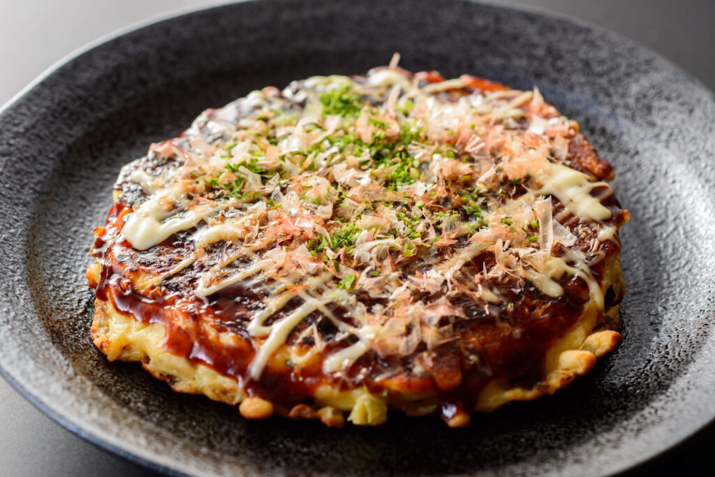 A black textured ceramic plate with one Osaka-style (Kansai-style) okonomiyaki on it. The pancake is topped with okonomiyaki sauce, mayonnaise, katsuobushi (bonito flakes) and green aonori seaweed.