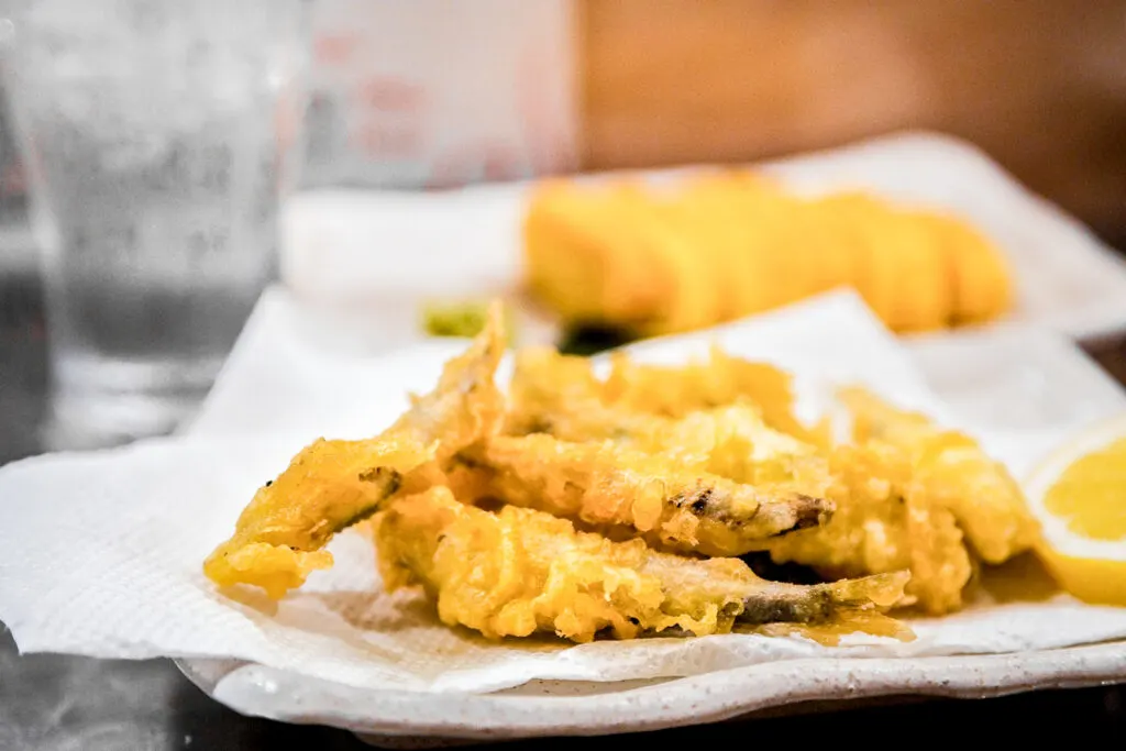 A serving of ayu tempura on a paper towel atop a white ceramic plate with a wedge of lemon. The ayu fish are small and whole, and have been battered and fried in tempura batter giving them a golden appearance.