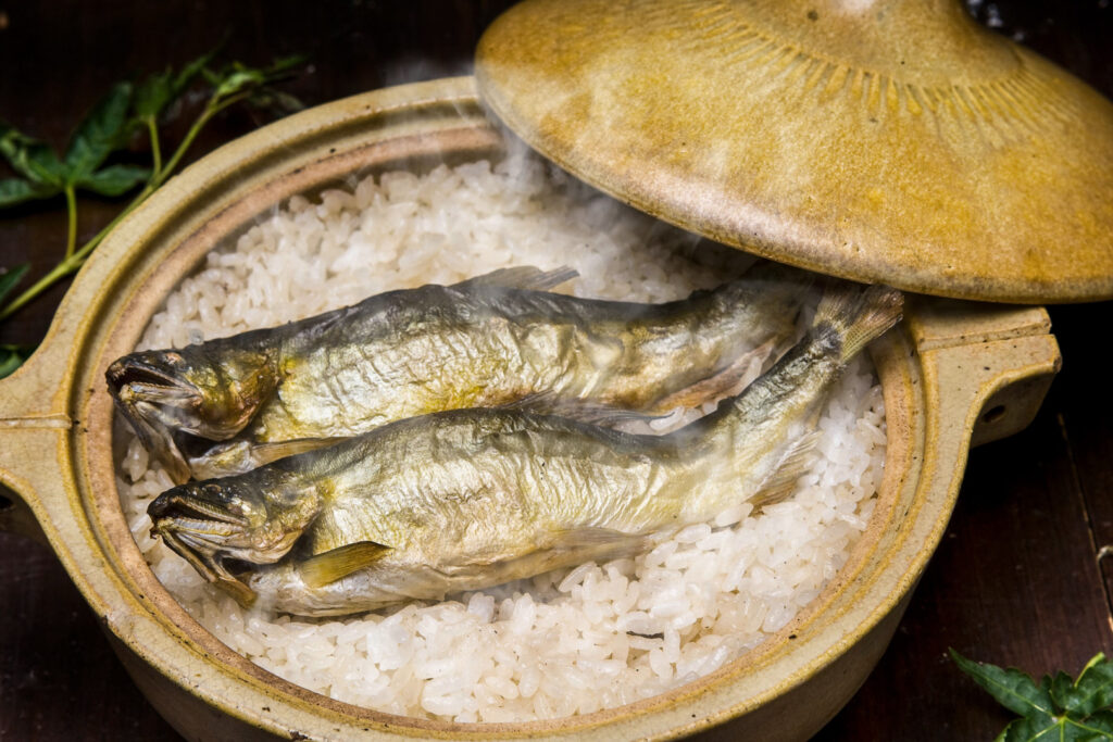 The lid of a yellow-mustard colored clay pot has been removed and is resting on the side of the pot. Inside we can see two whole ayu fish arranged on top of white rice. Steam can be seen rising up from the dish.