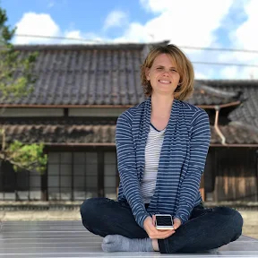 A photo of Japanese Food Guide contributor Jennifer Pastore. She is smiling and sitting cross-legged in front of a traditional Japanese building. Her hair is blonde and cut into a bob just above shoulder length. She is wearing a navy blue and white horizontal striped cardigan over a white v-neck top with fine horizontal blue stripes, dark pants, gray socks (no shoes) and holding a mobile phone in her hands. The sky above the building is blue with some fluffy white clouds.