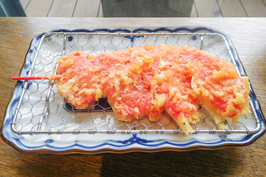 Osaka food: A large piece of red pickled ginger tempura (beni shoga ten) sits on a metal wire rack atop a blue and white ceramic plate on a wooden countertop. 