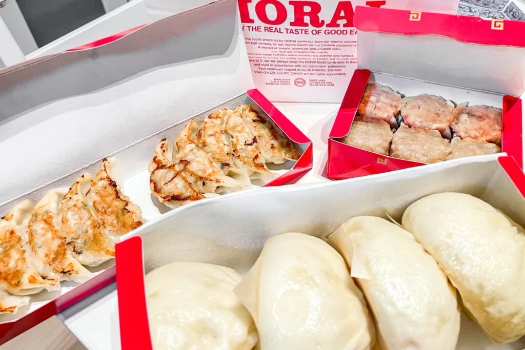 Osaka food: A paper bag from 551 Horai stands at the back with three open boxes in front. One contains four butaman, another ten gyoza and the other six shumai dumplings.