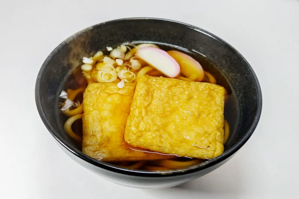 Osaka food: Kitsune udon in a black ceramic bowl. The dashi broth is brown in color and in it there is udon noodles, two large aburaage fried tofu pouches, two slices of pink and white kamaboko fish cake and some sliced up scallions or spring onions (the white part). 