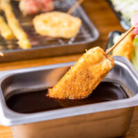 Osaka food: A hand can be seen dipping a skewered kushikatsu into a stainless steel rectangular metal container from the right of frame. What this particular kushikatsu contains is not exactly clear but could be cheese or a white fish. Other cooked kushikatsu ready for dipping can be seen in the background such as renkon lotus root and quail eggs.