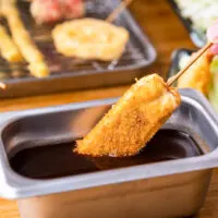 Osaka food: A hand can be seen dipping a skewered kushikatsu into a stainless steel rectangular metal container from the right of frame. What this particular kushikatsu contains is not exactly clear but could be cheese or a white fish. Other cooked kushikatsu ready for dipping can be seen in the background such as renkon lotus root and quail eggs.