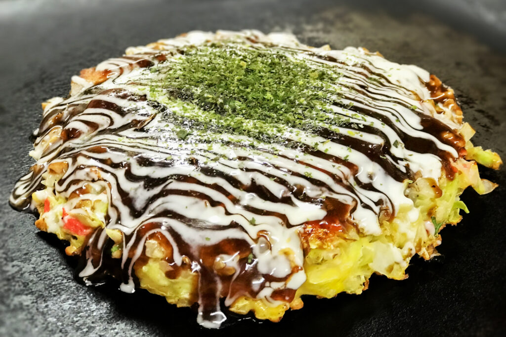 Osaka food: A Kansai-style okonomiyaki on a hotplate ready to eat. It has been topped with okonomiyaki sauce, mayonnaise and aonori seaweed.