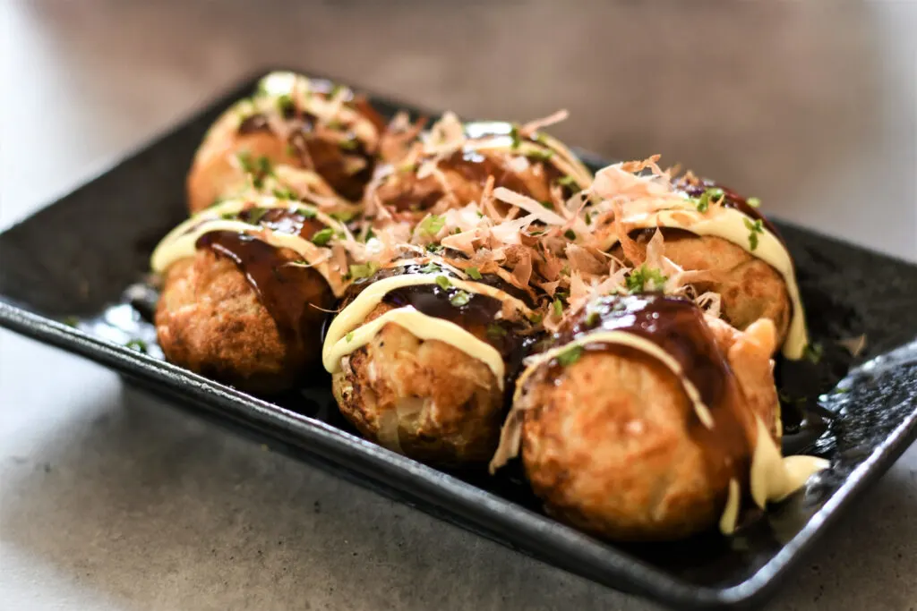 Osaka food: A serving of six takoyaki balls on a black rectangular ceramic plate. They have been topped with takoyaki sauce, mayonnaise, aonori seaweed and bonito flakes.