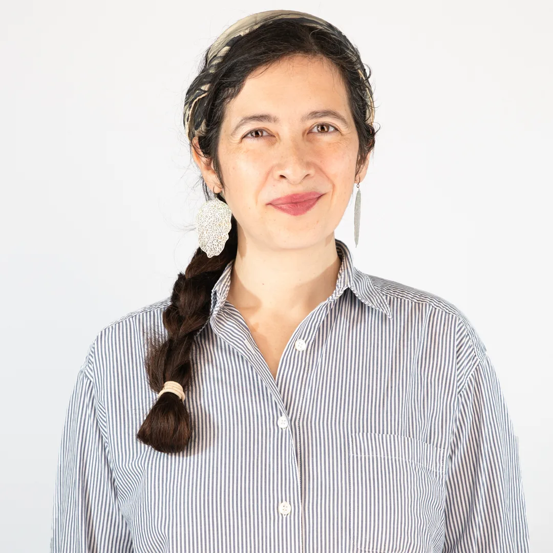 A photo of Japanese Food Guide contributor Selena Takigawa Hoy. She is facing the camera with a closed-mouth smile. She wears a white and blue striped shirt with collar, a blue/black and white headband, dangling silver earrings and her long, dark hair in a braid pulled over her right shoulder.