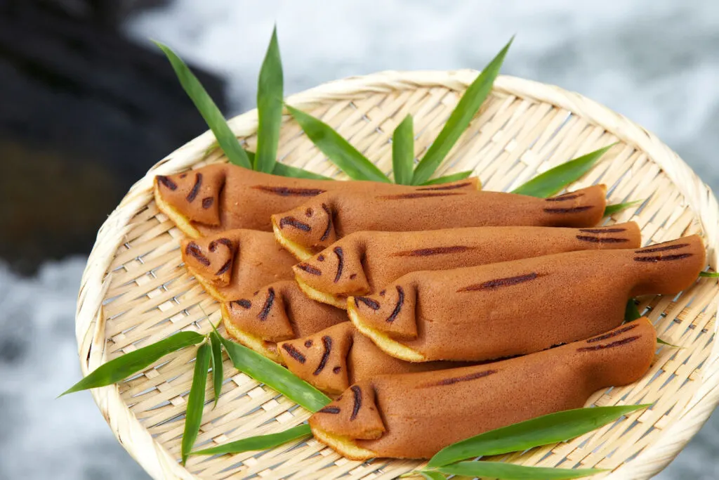 Eight ayu-gashi or waka-ayu castella cake sweets in the shape of ayu fish are arranged on a bamboo basket with some green foliage for decoration. The basket is being held in front of a river, the natural habitat of the ayu sweetfish.
