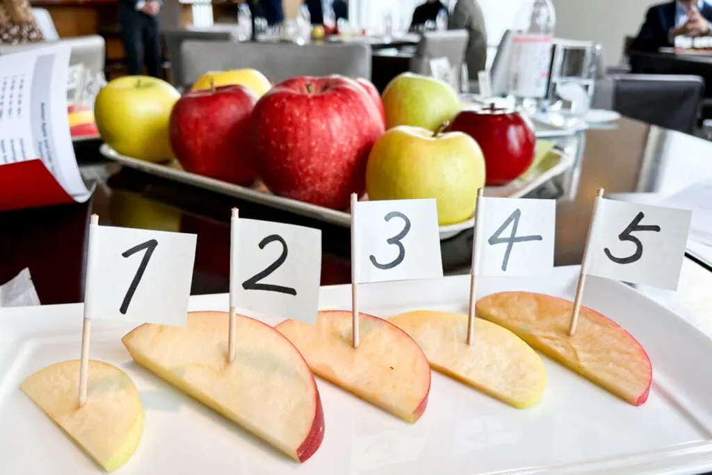 Aomori apple tasting: Five slices of apple on a rectangular white platter, each with a tooth pick inserted into it and a little white paper flag attached with numbers 1-5 written in black marker. Behind the apples for tasting is a platter of whole apples of different varieties.
