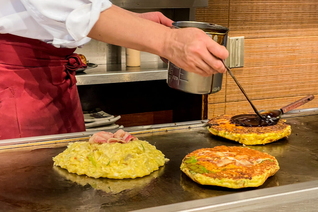 The torso and hands of the chef can be seen pouring brown okonomiyaki sauce from a metal container onto one of the three okonomiyaki cooking on the hot plate. The chef is wearing a white shirt and a red apron.