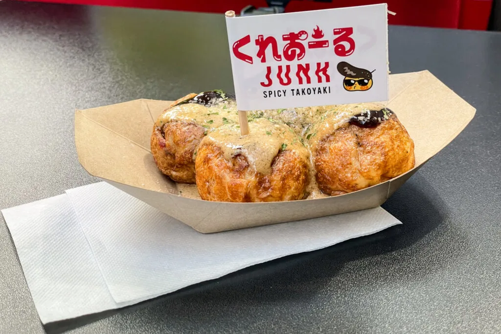 A serving of four porcini mushroom takoyaki with a toothpick flag poked into one of them that says "Creole JUNK" (the Creole part written in hiragana) and under it the words "Spicy takoyaki" in English. Next to the text is a little takoyaki character with a black hat and black sunglasses.