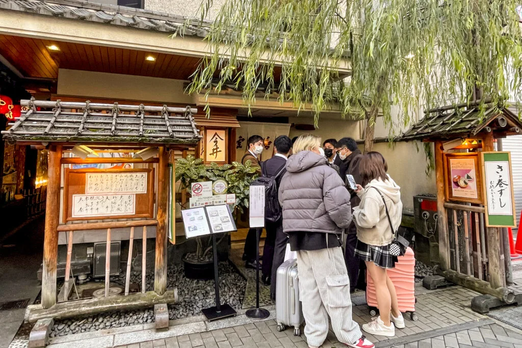 A line of people wearing face masks outside the traditional facade of the Dotonbori Imai main branch. A willow tree, that has become a landmark on Dotonbori can be seen out the front.
