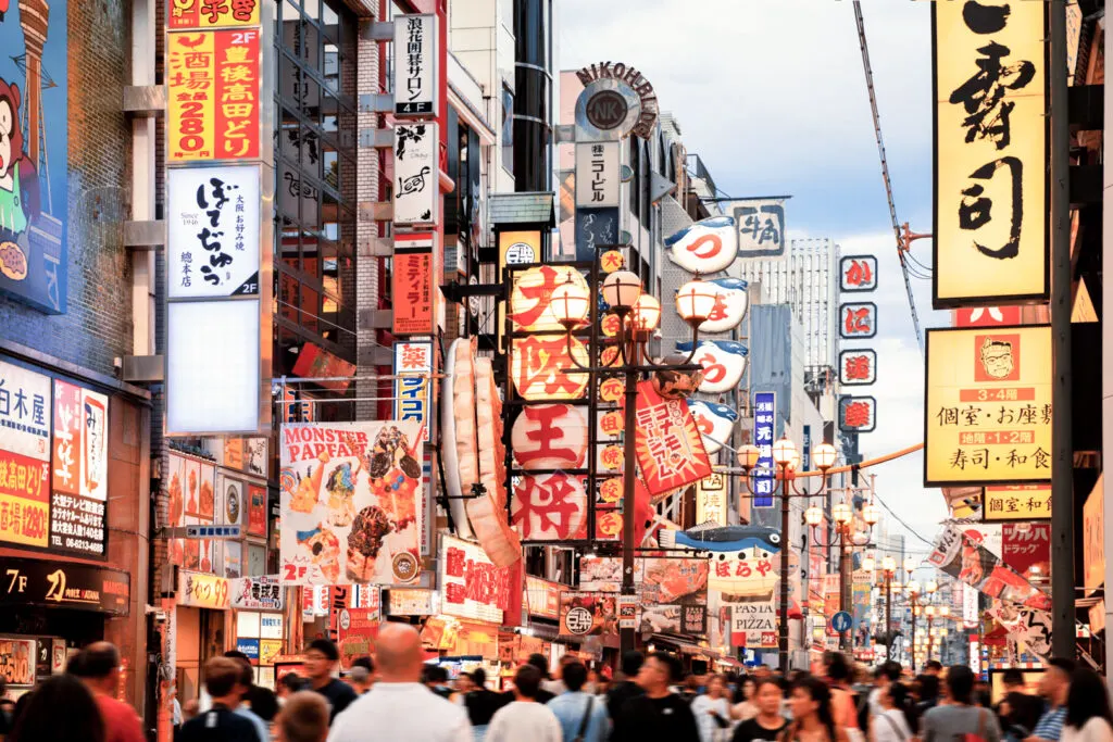 Looking down Dotonbori food street in Osaka. It's approaching dusk and the street is full of people walking in both directions. On either side of the street, the buildings feature many colorful and gaudy lit-up signs that have already been turned on in the fading light.