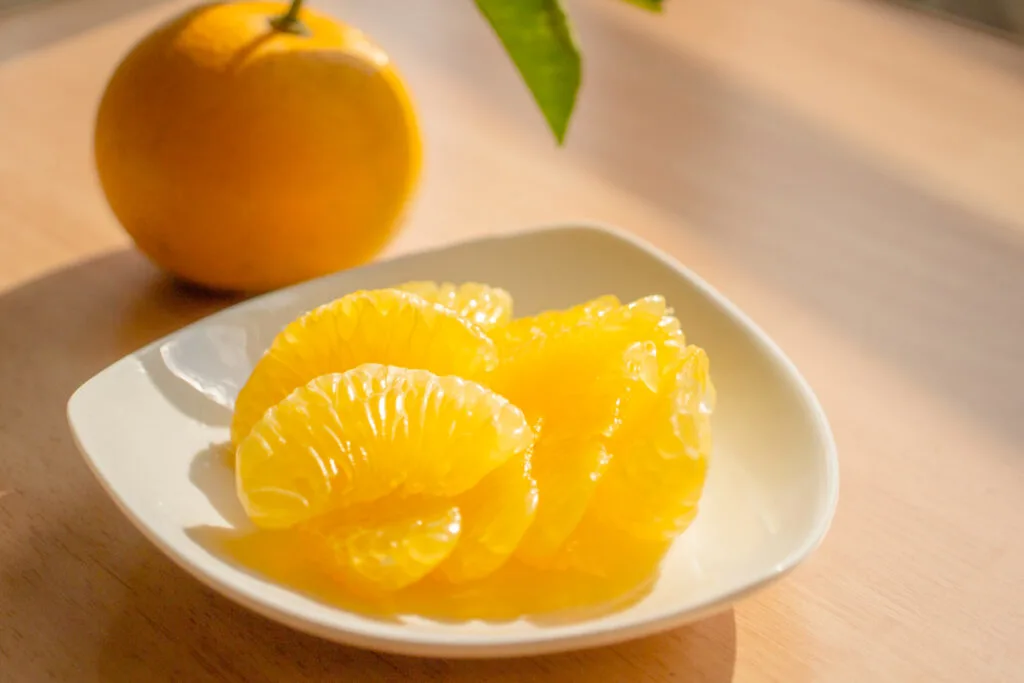 An orange hassaku citrus fruit with the peel on sits behind a small white ceramic plate with peeled segments of the yellowy-orange fruit arranged on it.
