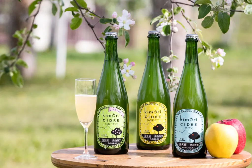 Japanese cider: Three bottles of Kimori Cidre sit on a table among the blossoms at Hirosaki Apple Park, where Kimori has an on-site cidery. From left to right is Kimori "Green" cidre (with a green label), then "Sweet" (yellow label), followed by "Dry" (blue label). To the left of the arrangement is some cider in a champagne flute and to the right two apples (one gold, one red).