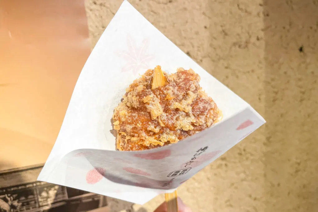 A fried momiji manju ('age momiji manju') being held out by the photographer in front of the camera on a wooden stick with a paper cone around the fried snack.